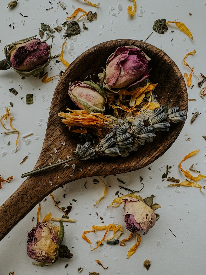 Whole herbs on a wooden spoon with scattered dried flowers. Herbs include lavender, rose and calendula, all known for healing the new mom after labor