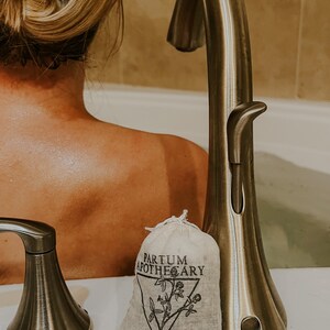 May include: A white muslin bag with black text that reads "Partum Apothecary" and a black illustration of a plant. The bag is sitting on a white surface next to a silver faucet.
