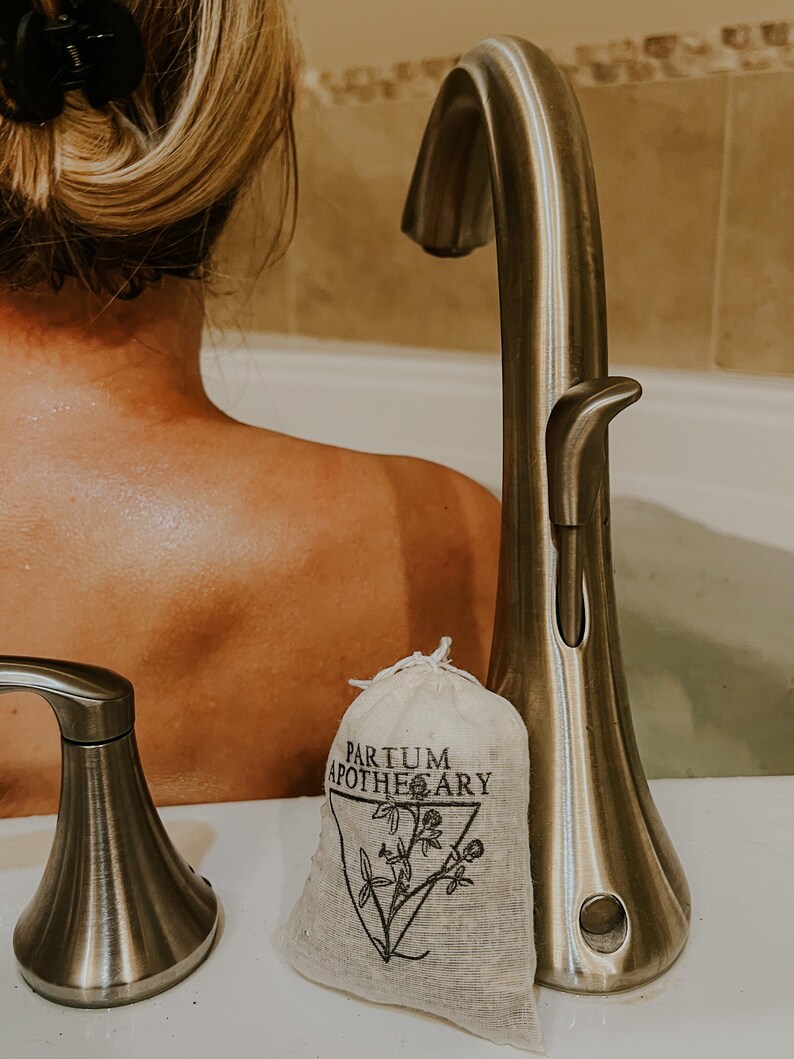 May include: A white muslin bag with black text that reads "Partum Apothecary" and a black illustration of a plant. The bag is sitting on a white surface next to a silver faucet.
