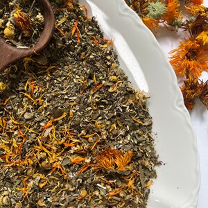 May include: A white ceramic bowl filled with a blend of dried herbs and flowers, including green leaves, yellow petals, and orange flower buds. The bowl is on a white surface.