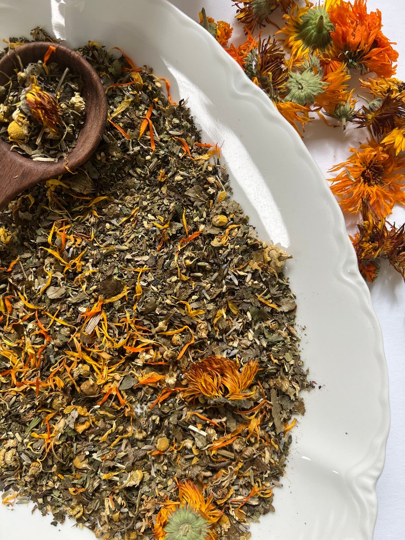 May include: A white ceramic bowl filled with a blend of dried herbs and flowers, including green leaves, yellow petals, and orange flower buds. The bowl is on a white surface.