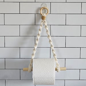 May include: A white paper towel holder made of macrame rope with a wooden dowel. The holder is hanging from a white tile wall.