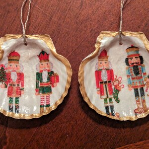 May include: Two Christmas ornaments made from seashells with a gold trim. Each ornament features a painted image of a nutcracker. One ornament has a red and green nutcracker, and the other has a blue and gold nutcracker.