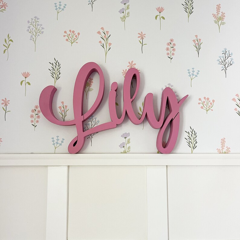 May include: Pink wooden letters spelling out the name "Lily" against a white background with a floral pattern.