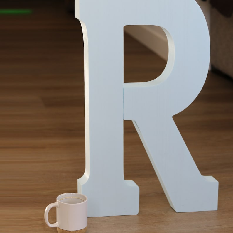 May include: A large, light blue wooden letter R stands on a wood floor. A small white ceramic mug sits in front of the letter.