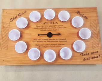 May include: Wooden shot glass roulette game with 12 white plastic shot glasses. The game board has the words "Shot Glass" and "Roulette" engraved on it, along with game rules and the phrase "Take your best shot!"