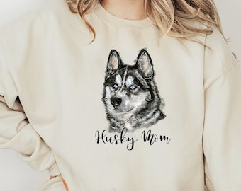 Op de afbeelding: Een crèmekleurige trui met een zwart-witte husky-hond illustratie en de tekst "Husky Mom" gedrukt op de voorkant.