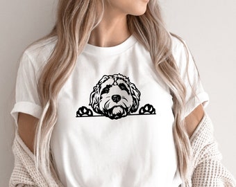 Puede incluir: Una camiseta blanca con una silueta negra de un perro asomándose por la parte superior. El perro tiene un pelaje esponjoso y mira a la cámara.
