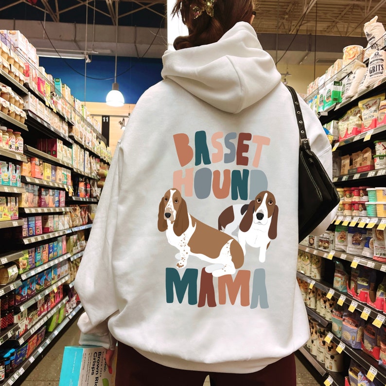 Pu&ograve; includere: Felpa con cappuccio bianca con un design grafico colorato di due Basset Hound e il testo "Basset Hound Mama".