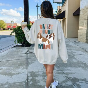 Pu&ograve; includere: Una felpa color crema con una grafica di due basset hound e il testo "Basset Hound Mama" in lettere colorate.