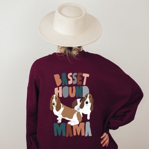 Pu&ograve; includere: Una felpa color borgogna con un design grafico di due basset hound e il testo "Basset Hound Mama" in lettere colorate e stilizzate.