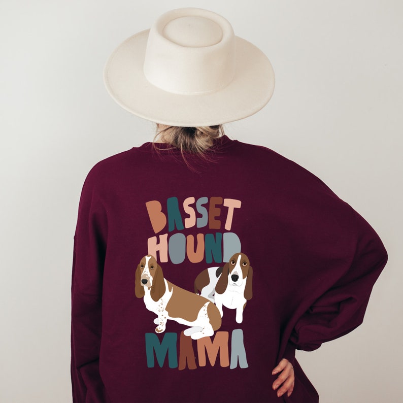 Pu&ograve; includere: Una felpa color borgogna con un design grafico di due basset hound e il testo "Basset Hound Mama" in lettere colorate e stilizzate.
