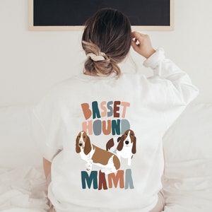 Pu&ograve; includere: Felpa bianca con una grafica colorata di due Basset Hound e il testo "Basset Hound Mama".