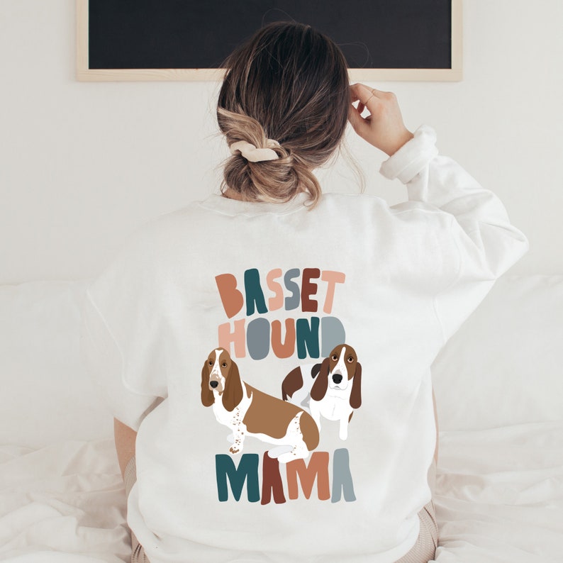 Pu&ograve; includere: Felpa bianca con una grafica colorata di due Basset Hound e il testo "Basset Hound Mama".
