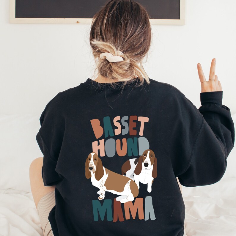 Pu&ograve; includere: Felpa nera con un design grafico di due basset hound e il testo "Basset Hound Mama" in lettere colorate in stile retr&ograve;.