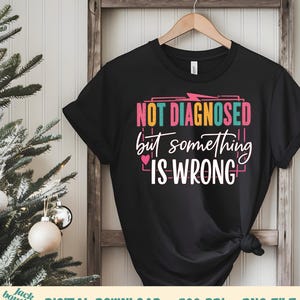 Op de afbeelding: Zwart t-shirt met de tekst "NOT DIAGNOSED but something IS WRONG" in roze, wit en teal. Het shirt hangt aan een houten hanger tegen een witte muur met een kerstboom op de achtergrond.