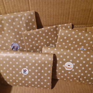 Puede incluir: Varios paquetes envueltos en papel marrón con un patrón de estrellas blancas están colocados dentro de una caja de cartón. Cada paquete está sellado con una pegatina redonda con varios personajes y diseños de dibujos animados. Listos para enviar.