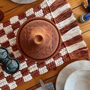 May include: A brown and white checkered table runner with fringe, a terracotta tagine pot with a lid, two blue ceramic salt and pepper shakers, and two white ceramic bowls with a white plate underneath each bowl.