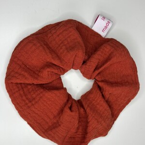 Peut inclure: Un grand chouchou de couleur rouille, fait de tissu texturé. Le chouchou est circulaire et porte une étiquette avec l'inscription "made ja". Cet accessoire pour cheveux est conçu pour maintenir les cheveux en queue de cheval ou en chignon.