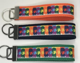 Pode incluir: Três chaveiros com um design dos Beatles colorido. Os chaveiros são laranja, preto e verde. Cada chaveiro tem um aro de chave prateado.