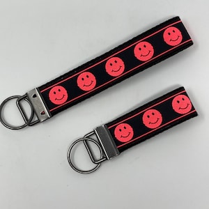 May include: Two black keychains with red smiley faces. The larger keychain has five smiley faces and the smaller keychain has three smiley faces.