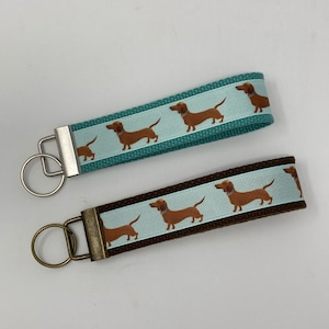 May include: Two dachshund dog keychains. One keychain has a teal blue strap with a white stripe and brown dachshund silhouettes. The other keychain has a brown strap with a white stripe and brown dachshund silhouettes.
