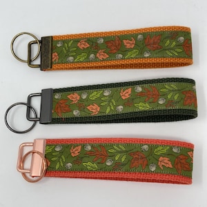 May include: Three keychains with fall-themed designs. Each keychain features a fabric strap with a green leaf and acorn pattern, bordered by orange, dark green, and coral-colored woven fabric. The key rings and hardware have different metallic finishes.