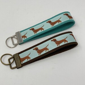 May include: Two dachshund dog keychains. One keychain has a teal blue strap with a white ribbon featuring brown dachshunds. The other keychain has a brown strap with a light blue ribbon featuring brown dachshunds.