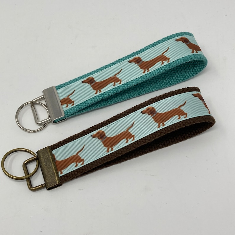 May include: Two dachshund dog keychains. One keychain has a teal blue strap with a white ribbon featuring brown dachshunds. The other keychain has a brown strap with a light blue ribbon featuring brown dachshunds.