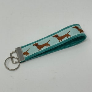 May include: Teal blue key chain with a white ribbon featuring brown dachshund silhouettes. The key chain has a silver metal key ring.