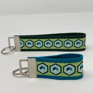 May include: Two key fobs with geometric patterns. One is dark green with a light green band featuring blue, teal, and white hexagons. The other is teal with a light green band and the same geometric design. Both have silver key rings.