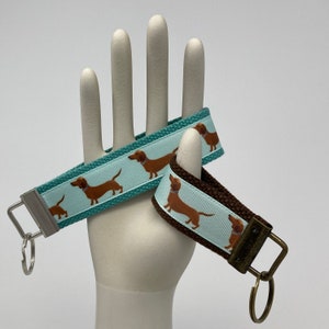May include: Two dachshund dog keychains, one with a teal blue background and the other with a brown background. Both keychains feature a white background with brown dachshund dogs printed on them.