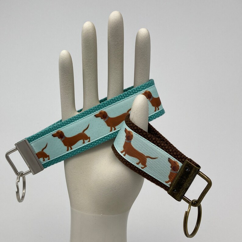 May include: Two dachshund dog keychains, one with a teal blue background and the other with a brown background. Both keychains feature a white background with brown dachshund dogs printed on them.