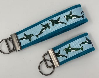 May include: Two teal blue keychains with silver key rings. The keychains feature a light blue ribbon with a repeating pattern of black and green glitter orca whales.