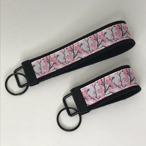 May include: Two black keychains with pink and white cherry blossom patterned fabric. The keychains are different lengths.