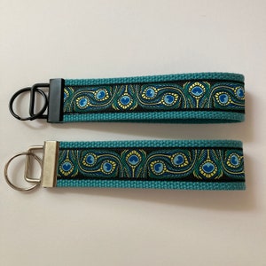 May include: Two teal keychains with a black and gold peacock feather design. The keychains have a black and silver metal ring. The keychains are made of a durable fabric.