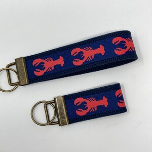 May include: Two navy blue keychains with red lobster patterns. The keychains have bronze-colored metal hardware. One keychain is larger than the other. The keychains are made of a woven fabric.