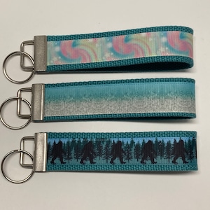 May include: Three teal blue keychains with silver key rings. The first keychain has a pink and blue swirl pattern. The second keychain has a light blue and white ombre pattern. The third keychain has a black silhouette of Bigfoot walking through a forest.