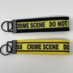 May include: Two keychains with black metal rings and black and yellow bands. The bands read "CRIME SCENE DO NOT CROSS" in black text. One keychain has a black band, and the other has a yellow band.
