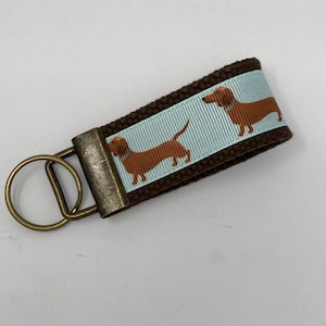 May include: Brown canvas keychain with a light blue ribbon featuring a dachshund print. The keychain has a brass key ring.