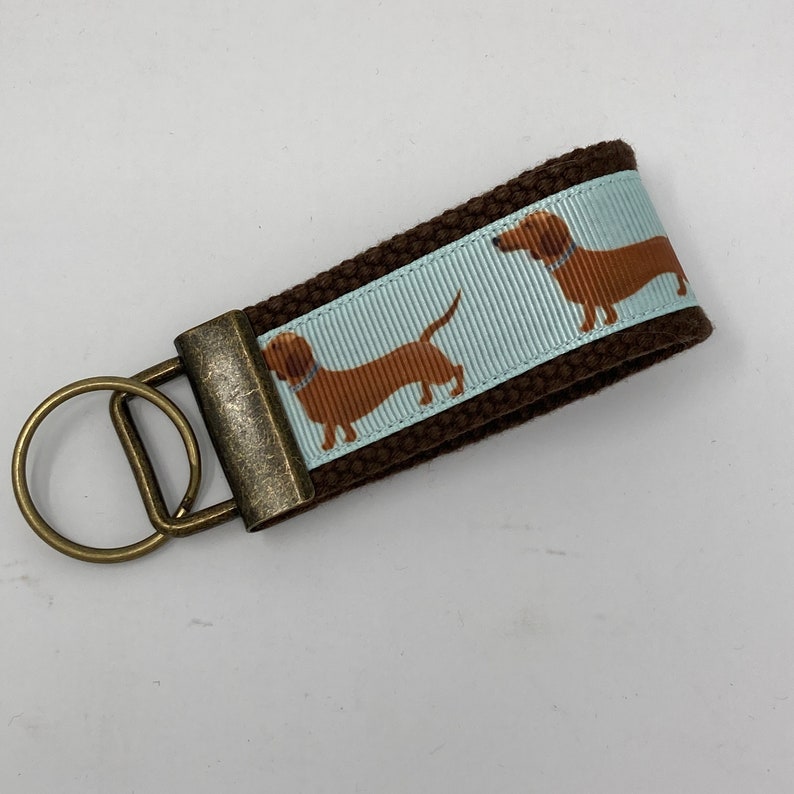 May include: Brown canvas keychain with a light blue ribbon featuring a dachshund print. The keychain has a brass key ring.