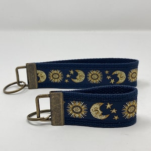 May include: Two navy blue keychains with gold embroidered suns, moons, and stars. The keychains have antique brass key rings.