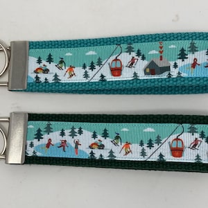 May include: Two keychains with a ski resort theme. One keychain is teal with a ski resort ribbon design, and the other is dark green. Both have a silver metal key ring.