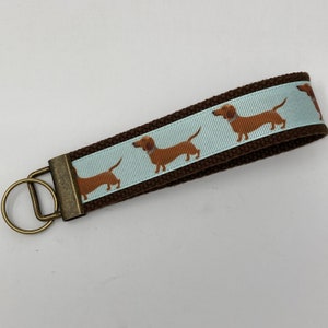 May include: Brown canvas keychain with a light blue ribbon featuring a repeating pattern of brown dachshund dogs. The keychain has a brass key ring.