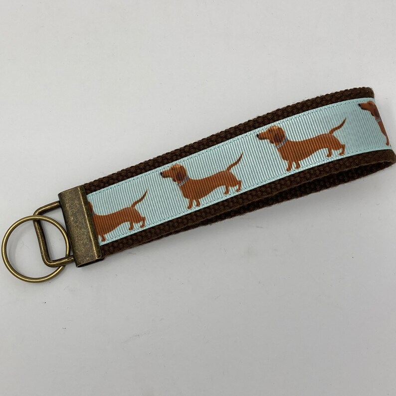 May include: Brown canvas keychain with a light blue ribbon featuring a repeating pattern of brown dachshund dogs. The keychain has a brass key ring.
