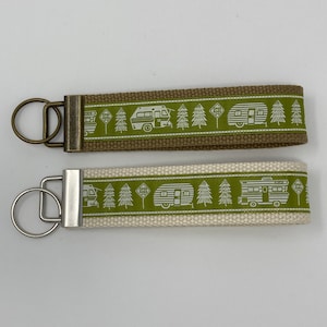 May include: Two keychains with a green and white pattern of a camper, pine trees, and a "park here" sign. The keychain on the top is on a brown strap and the keychain on the bottom is on a white strap.