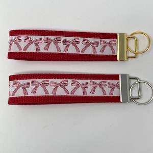 Puede incluir: Dos llaveros rojos con diseños de lazos blancos. Un llavero tiene un anillo de llave dorado y el otro tiene un anillo de llave plateado.