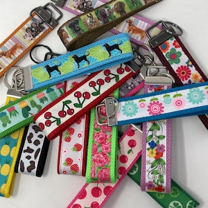 May include: A collection of fabric key fobs featuring various patterns including black dogs on blue, red cherries on white, pink roses, strawberries, colorful flowers, pineapples, and cow print. Each strap has a metal hardware clasp and ring.