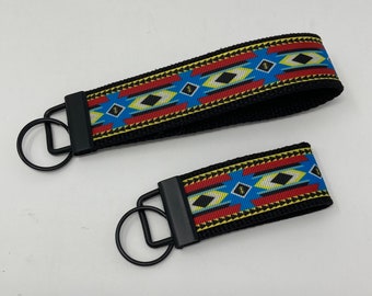 May include: Two black keychains with a colorful geometric pattern. The pattern features red, yellow, blue, and white stripes with black accents. The keychains are made of black fabric and have a metal ring.