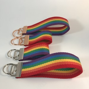 May include: Three rainbow striped keychains with silver and gold key rings. The keychains are made of woven fabric with red, orange, yellow, green, blue, indigo, and violet stripes.